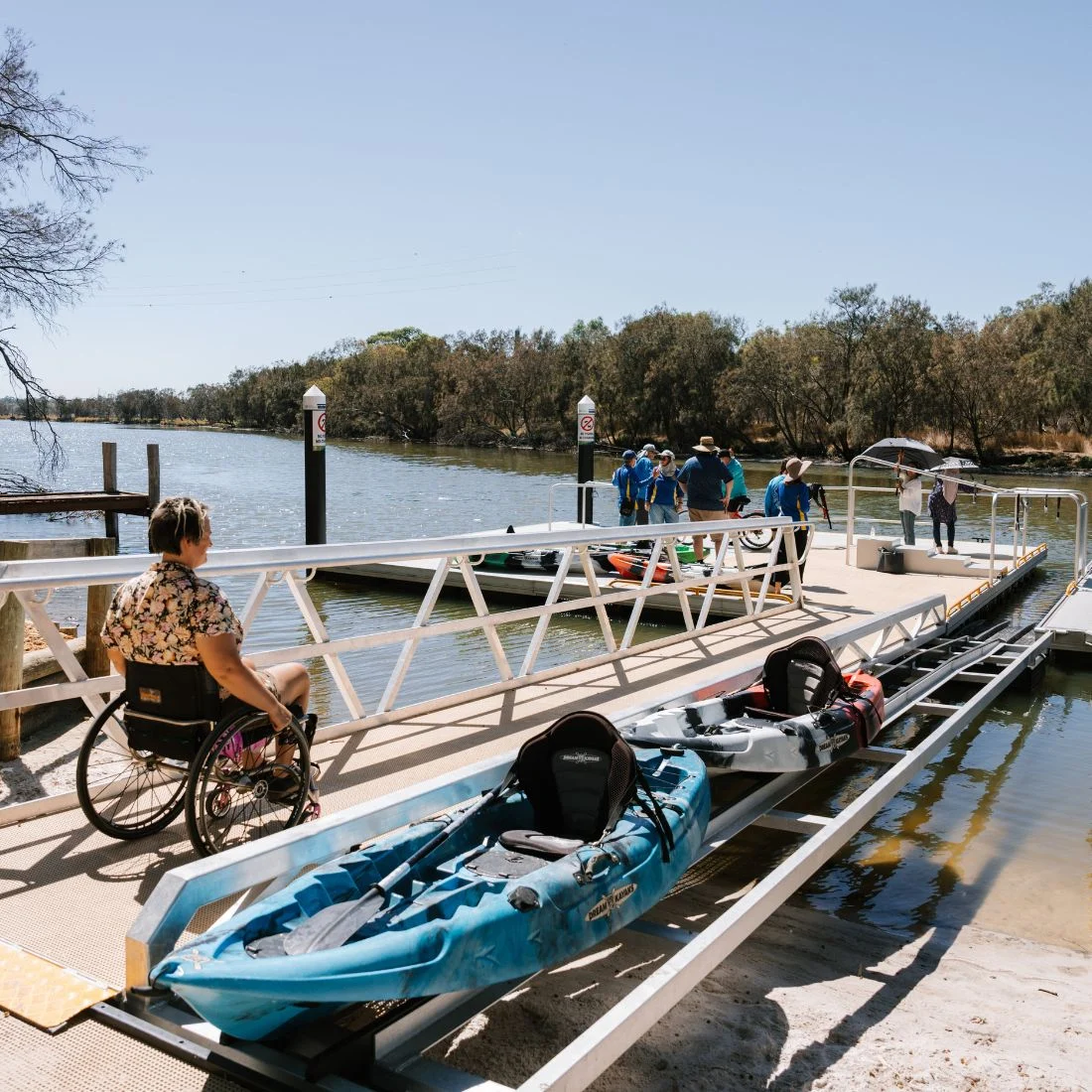 A person in a wheelchair waits on a ramp near a dock where people prepare kayaks on a sunny riverside. The dock has accessible features for wheelchairs and kayak launches. Trees line the water in the background.