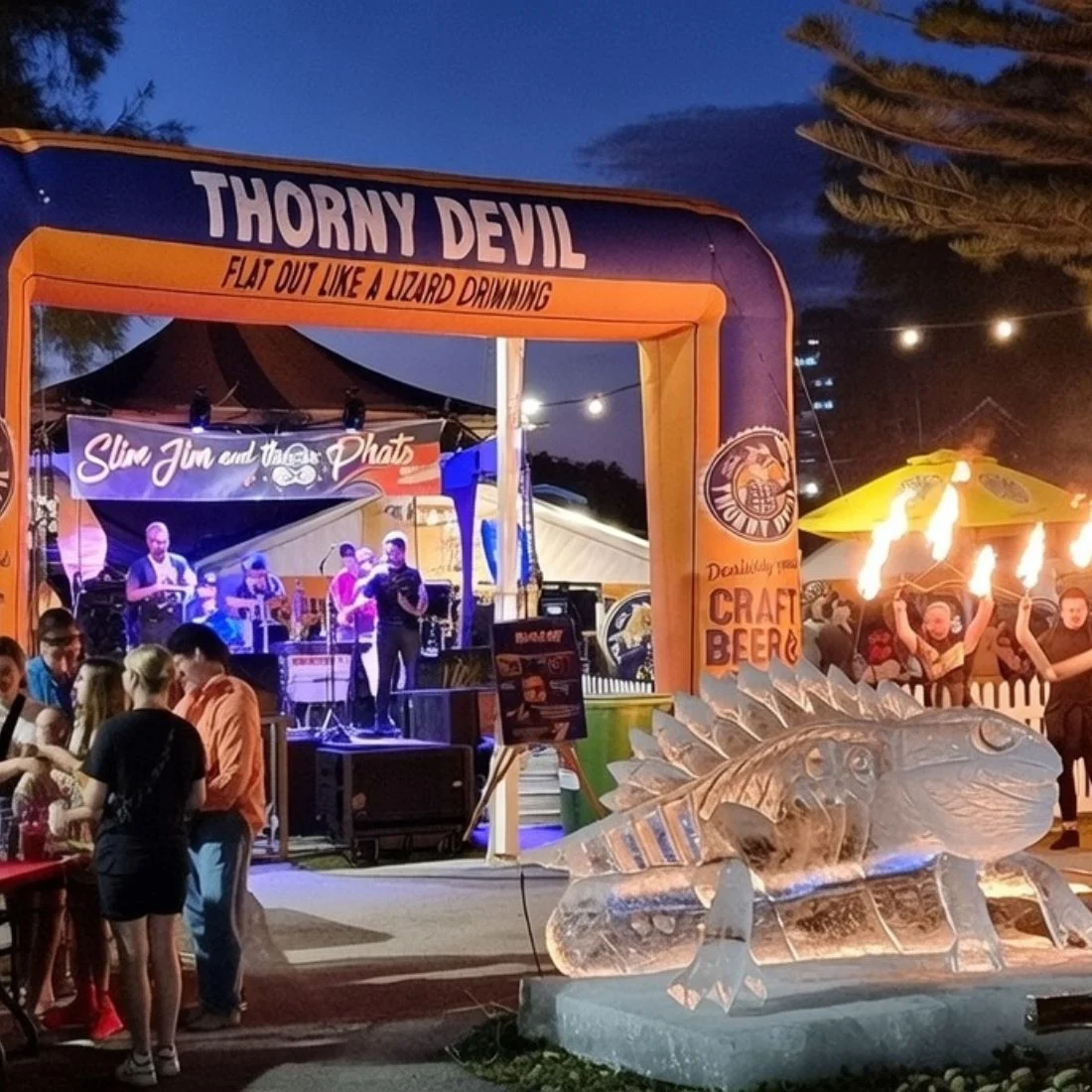 A lively outdoor event at dusk features a band performing on stage, people socializing, an illuminated Thorny Devil arch, and a detailed ice sculpture of a lizard. Bright lights and decorations enhance the festive atmosphere.