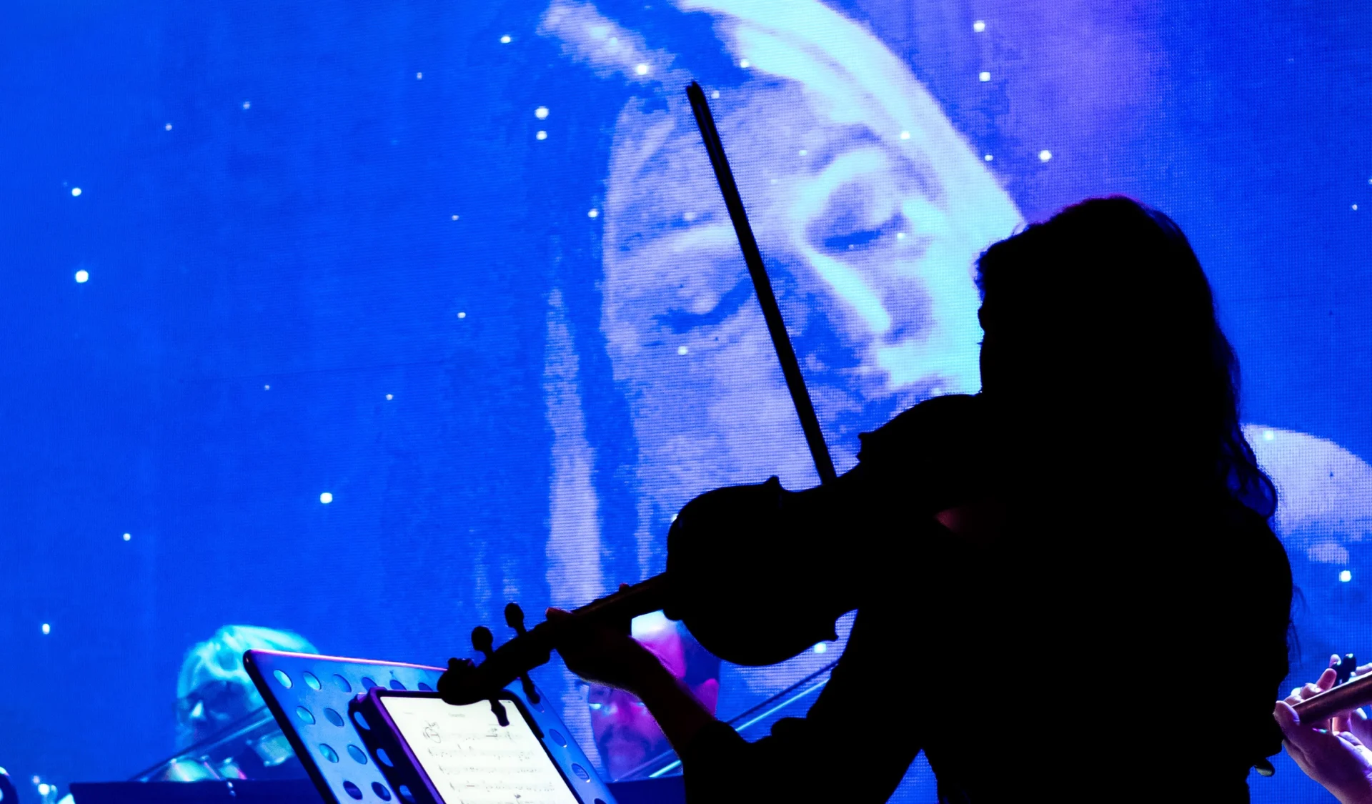 A silhouetted violinist plays in front of a large screen displaying a close-up of a womans face against a starry, blue background.