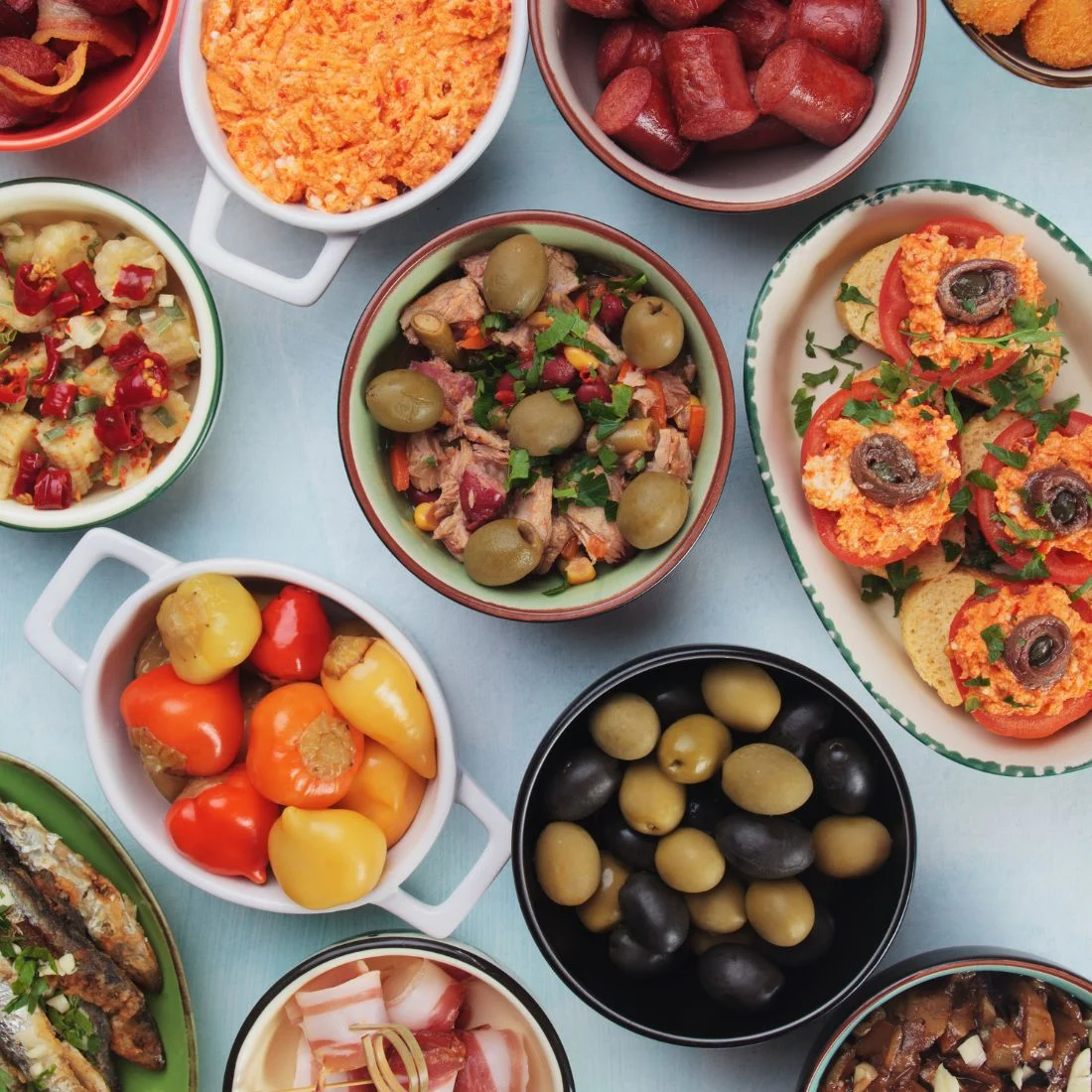 A spread of various tapas dishes including olives, peppers, sardines, and tomato appetizers.