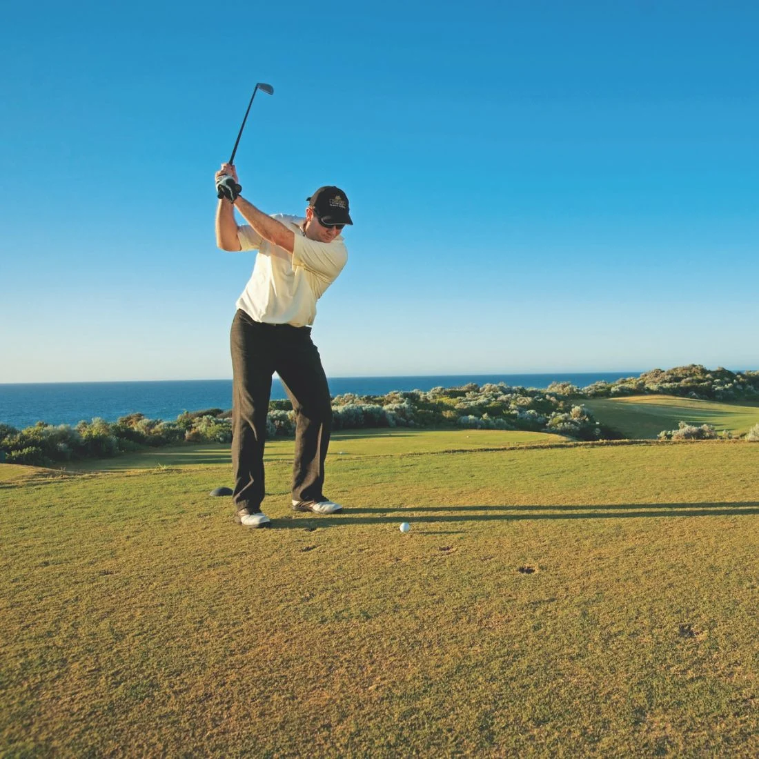 A person in a yellow shirt and dark pants swings a golf club on a grassy course with the ocean in the background under a clear blue sky.