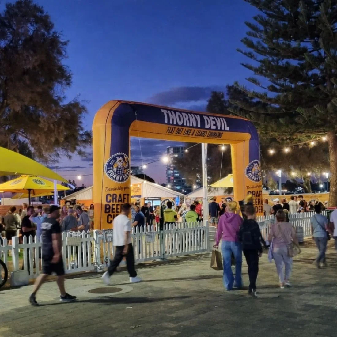A lively outdoor beer festival at dusk features crowds, yellow tents, and a large inflatable arch reading Thorny Devil. People stroll and mingle near white picket fences under string lights and tall trees.