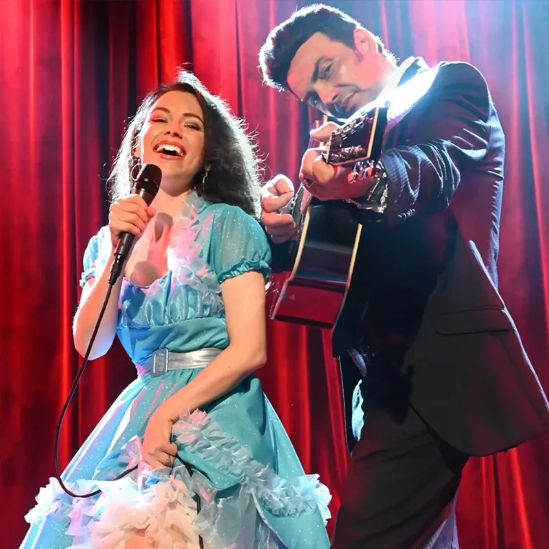 A woman in a sparkly blue dress sings into a microphone while a man in a black suit plays an acoustic guitar.