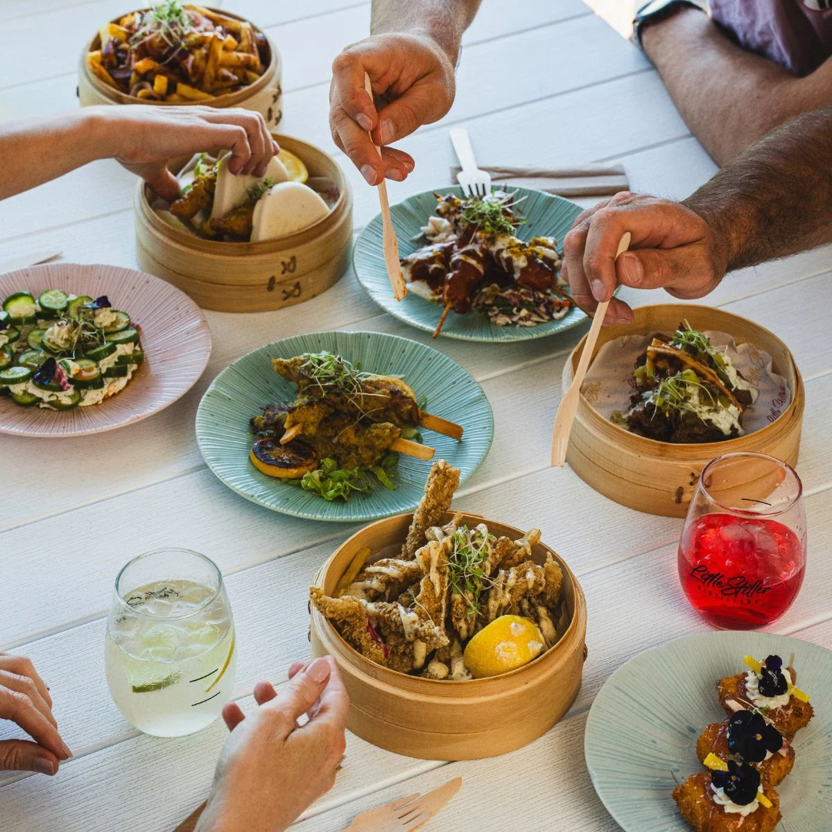 A table laden with various dishes, including skewers, fried seafood, fries, and small appetizers, with drinks and hands reaching for the food.