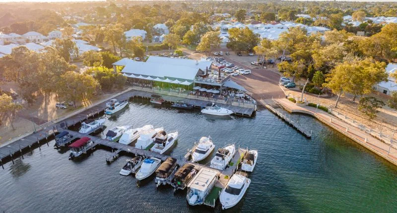 An aerial view of a marina with many boats docked, a large restaurant building with outdoor seating, and surrounding trees and houses under a golden sunset.