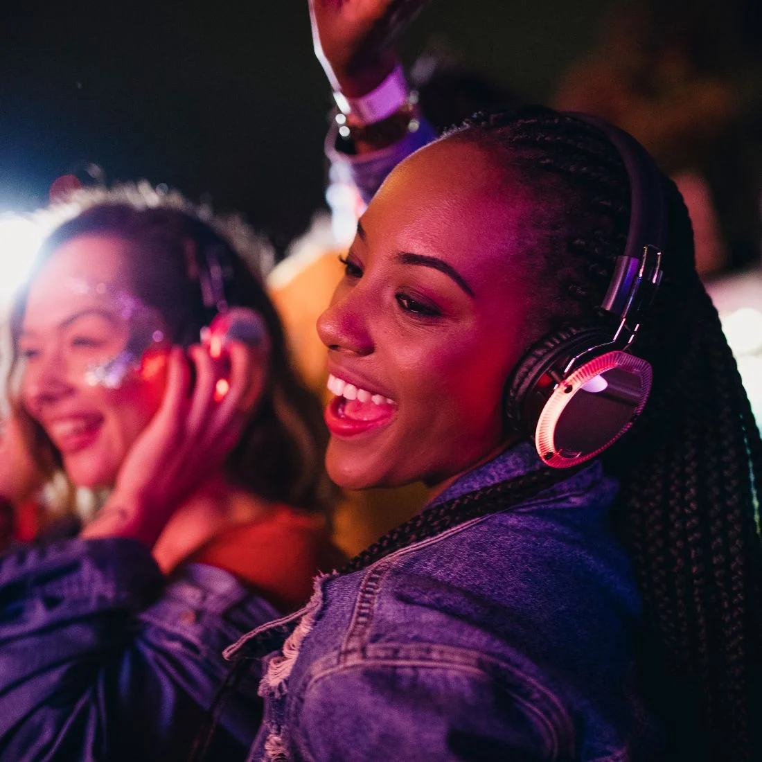 Two young women wearing headphones and enjoying music at an event with colorful lighting.