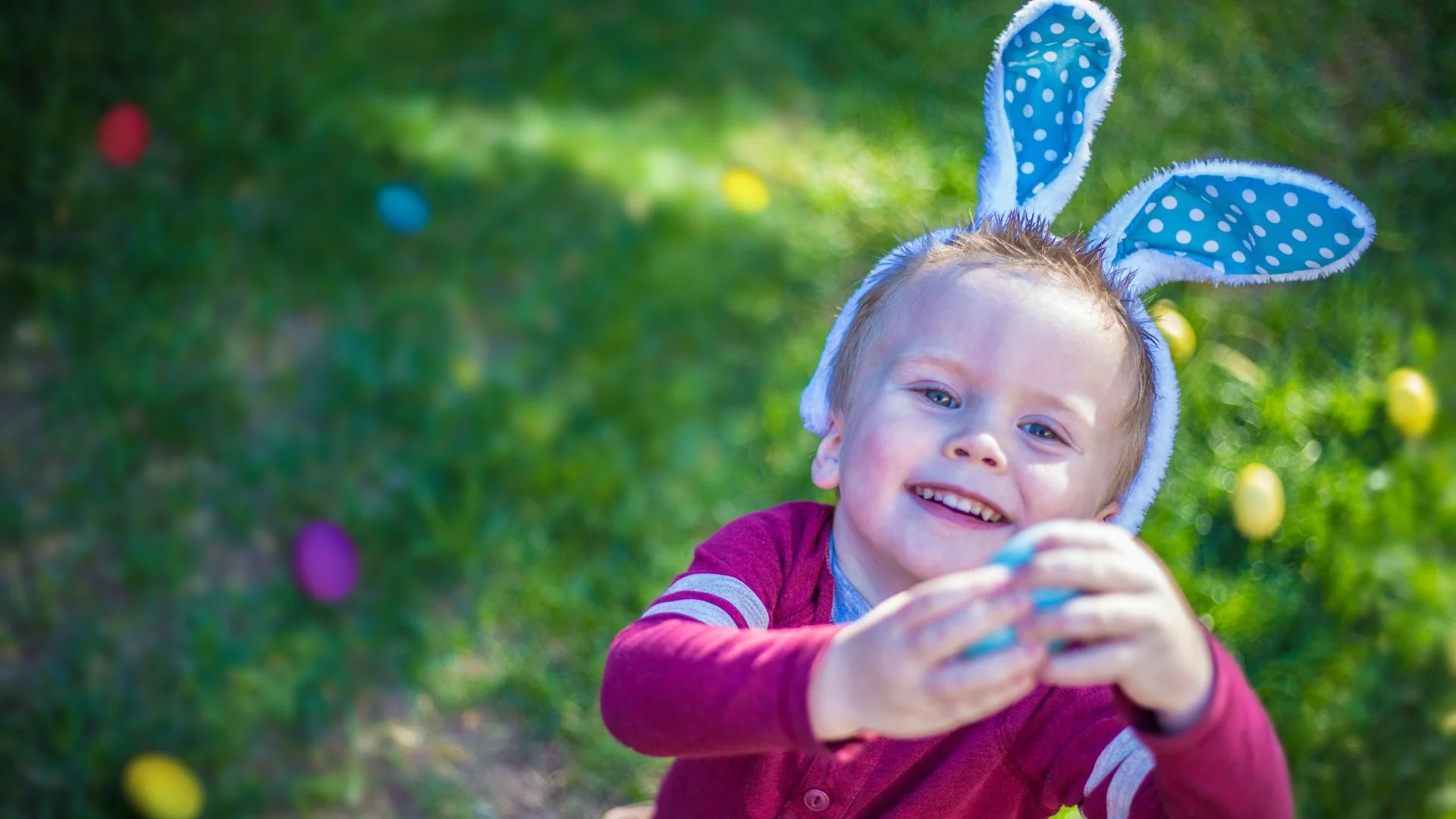 A child wearing blue and white polka dot bunny ears smiles while holding a blue Easter egg. Colorful Easter eggs are scattered on the green grass in the background.