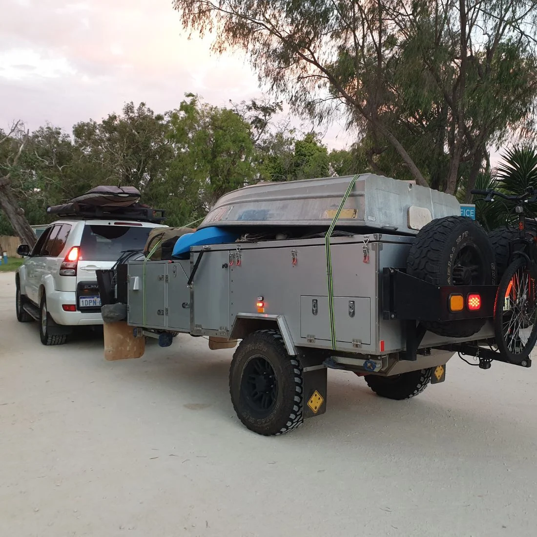 A white SUV is towing an off-road camper trailer loaded with a small boat, bicycles, and various gear. The scene is set on a dirt road, surrounded by trees and greenery, during late afternoon or early evening.