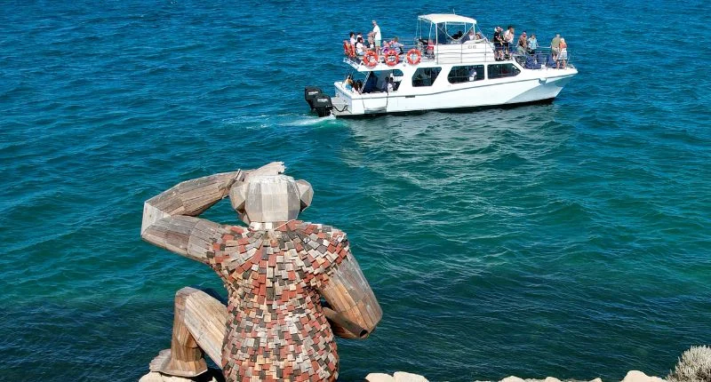 A Giant sculpture made of wood sits on a cliff overlooking the blue ocean, where a white tour boat with several passengers sails by.