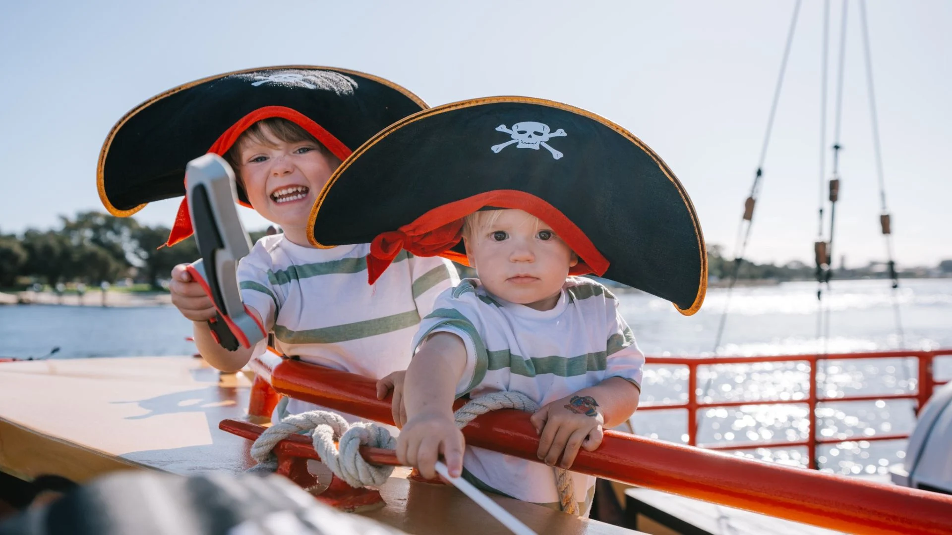 Two young children wearing pirate hats and striped shirts play on a boat. One child grins and holds a toy hook, while the other looks at the camera, holding a toy sword. Water and trees are visible in the background.