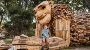 A young boy sits on the wooden hand of a large, whimsical wooden sculpture of an ape in an outdoor park surrounded by trees.