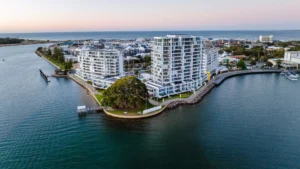 Aerial view of modern white apartment buildings on a small peninsula surrounded by water, with a marina and a town in the background under a clear sky at sunset.



