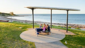A family of four enjoys a picnic at a table under a modern shelter near a beach, with the sea and shoreline in the background on a sunny day.