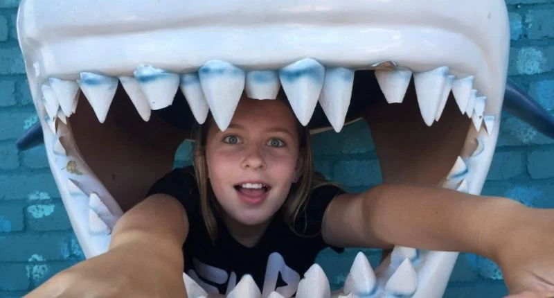 A girl peeks out from inside the open mouth of a shark prop with large, white teeth.