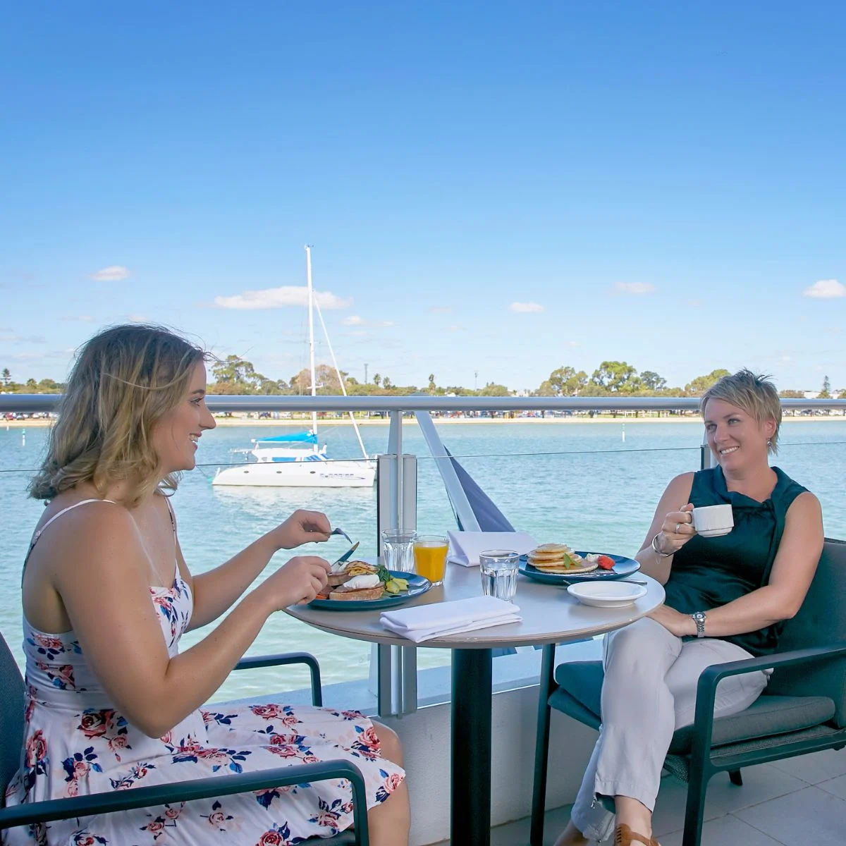 Two women enjoying breakfast at a table overlooking the Mandurah Estuary with a sailboat in the background.