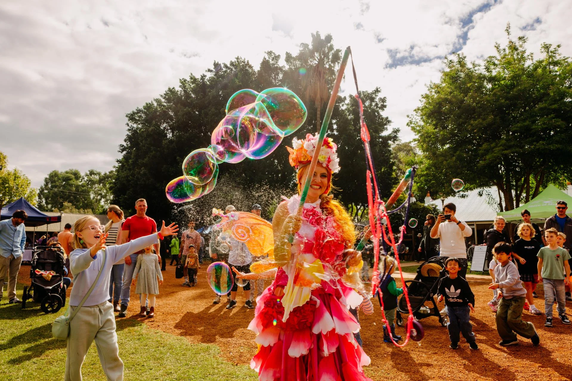 A performer in a colorful flower costume makes large bubbles with a wand at an outdoor festival as excited children and families watch and reach for the bubbles under a partly cloudy sky.