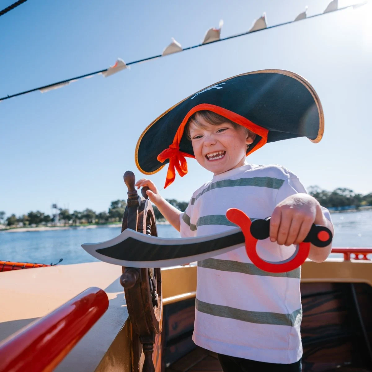 A young boy wearing a pirate hat and holding a toy sword stands at a ship's wheel on a sunny day.