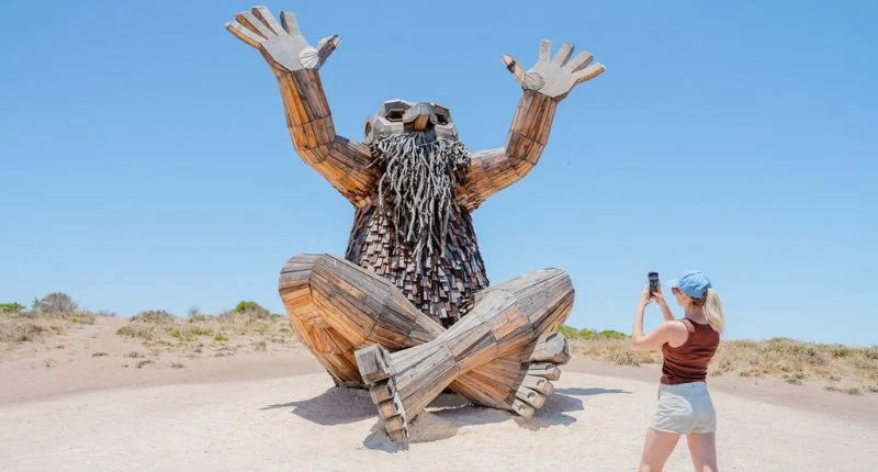 A large wooden sculpture of a seated figure with a long beard and raised arms, with a person in the foreground taking a photo of it with a smartphone.