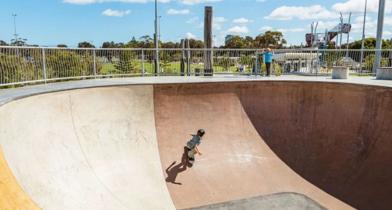 A young boy skateboarding in a large concrete skatepark on a sunny day.