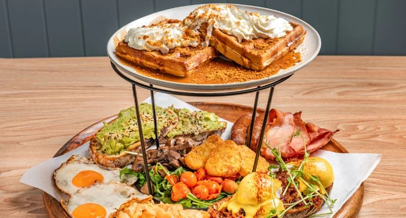 A two-tiered platter filled with a variety of breakfast foods including waffles with whipped cream, avocado toast, fried eggs, bacon, sausage, hash browns, and eggs benedict.