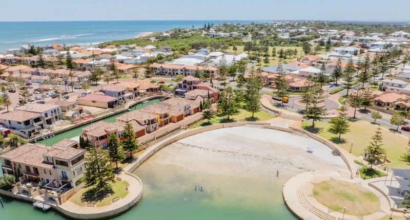 An aerial view of a coastal residential area with canals, houses, and a sandy beach area. The ocean is visible in the background under a clear blue sky.