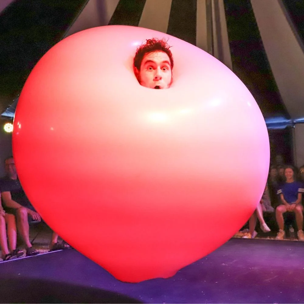 A performer with a surprised expression has their head sticking out of the top of a large, inflated pink balloon, surrounded by an audience under a circus tent.
