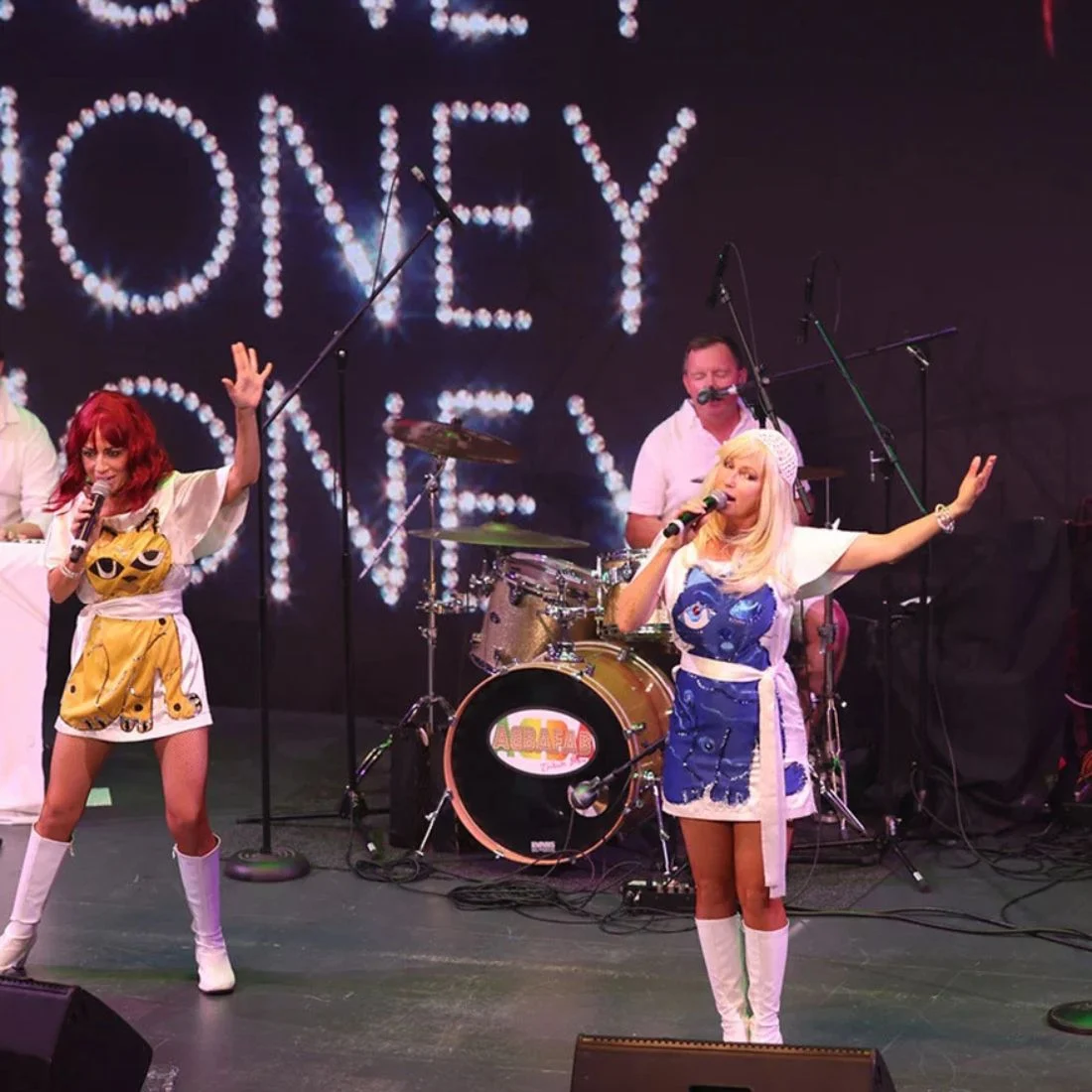 A musical duo dressed in cat-themed outfits performs on stage with a drummer in the background. The stage backdrop features the word "MONEY" spelled out in sparkling lights.