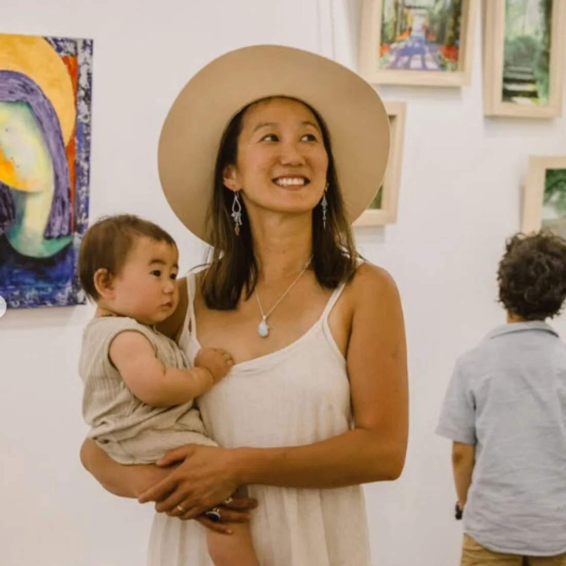 A woman in a light dress and wide-brimmed hat holds a baby while smiling at an art gallery. Colorful paintings hang on the walls and a person stands in the background viewing the art.
