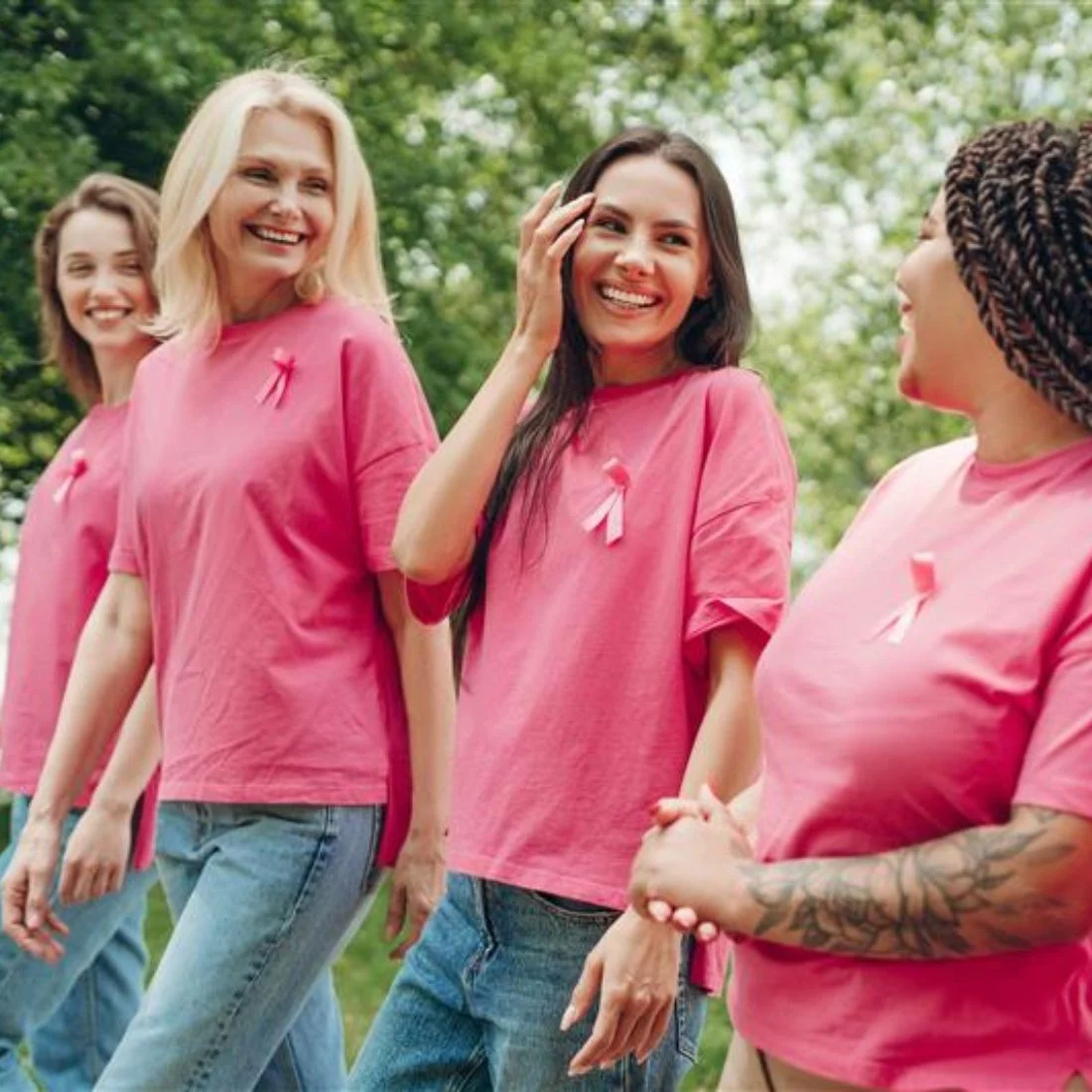 Four women wearing pink shirts with pink ribbons walk together outdoors, smiling and talking, in a show of support for breast cancer awareness.
