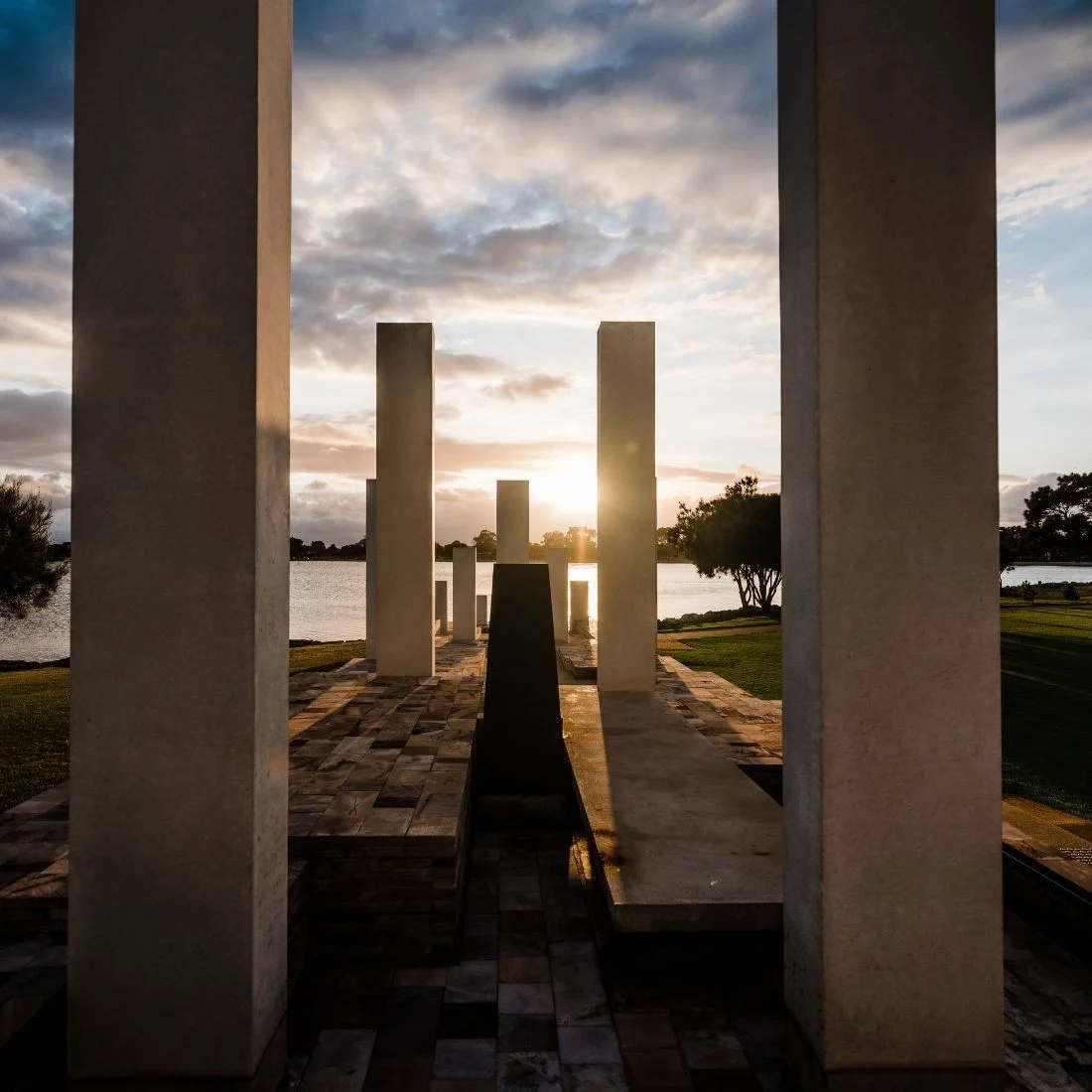 Tall rectangular stone pillars and low walls form a modern outdoor monument by a lake, with the sun setting in the background and clouds filling the sky. Trees and grass frame the peaceful scene.