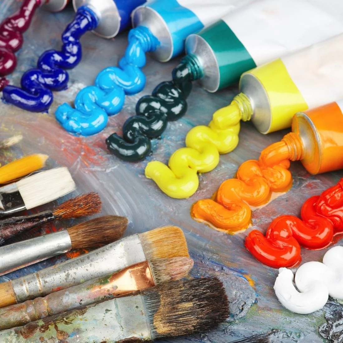 Tubes of colorful paint squeezed onto a palette with thick lines of paint in blue, green, yellow, orange, red, and white, surrounded by several used paintbrushes with paint on their bristles.