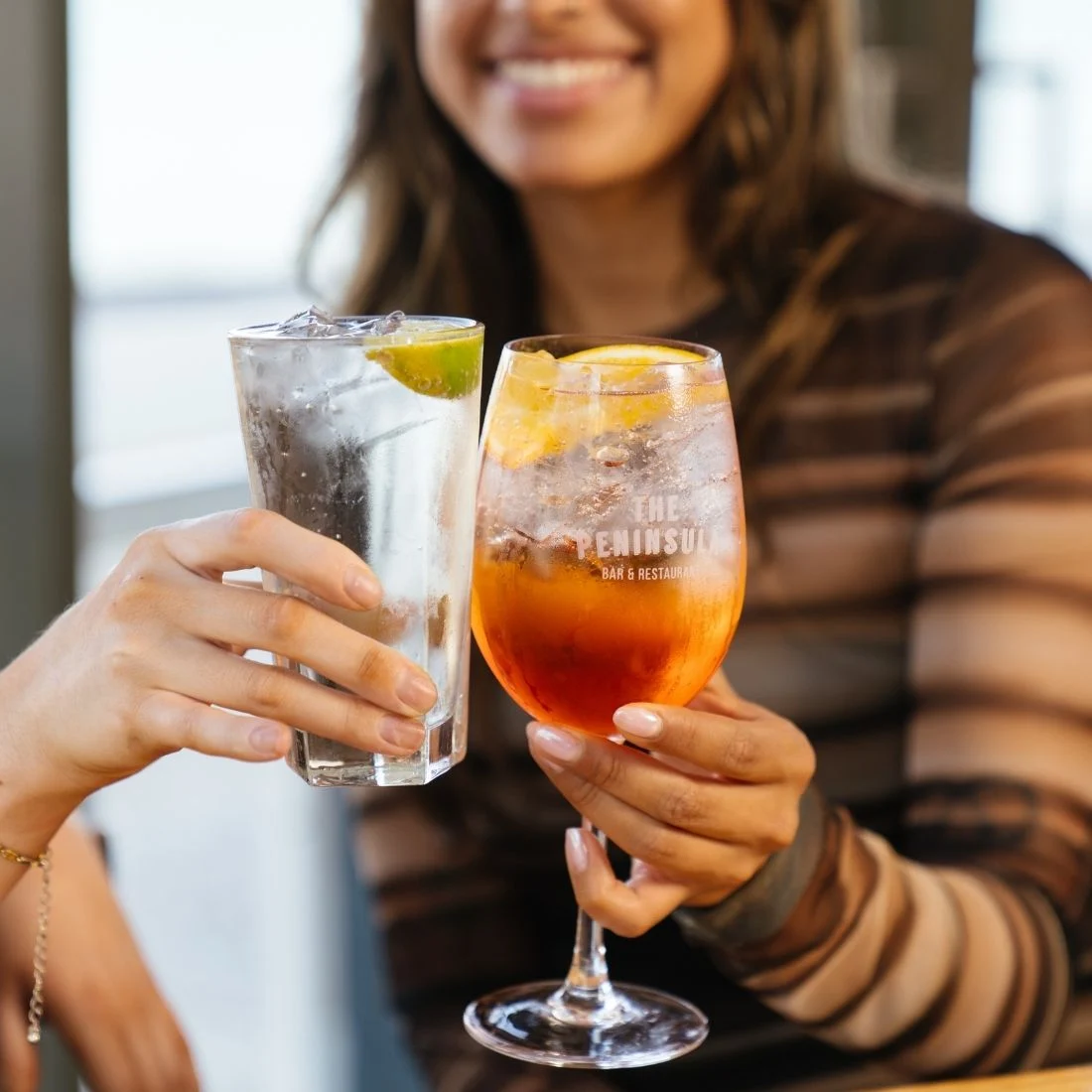 Two people cheersing drinks—one holding a clear cocktail with lime in a tall glass, the other holding an orange cocktail in a wine glass. Both are smiling, and the setting appears relaxed and social.