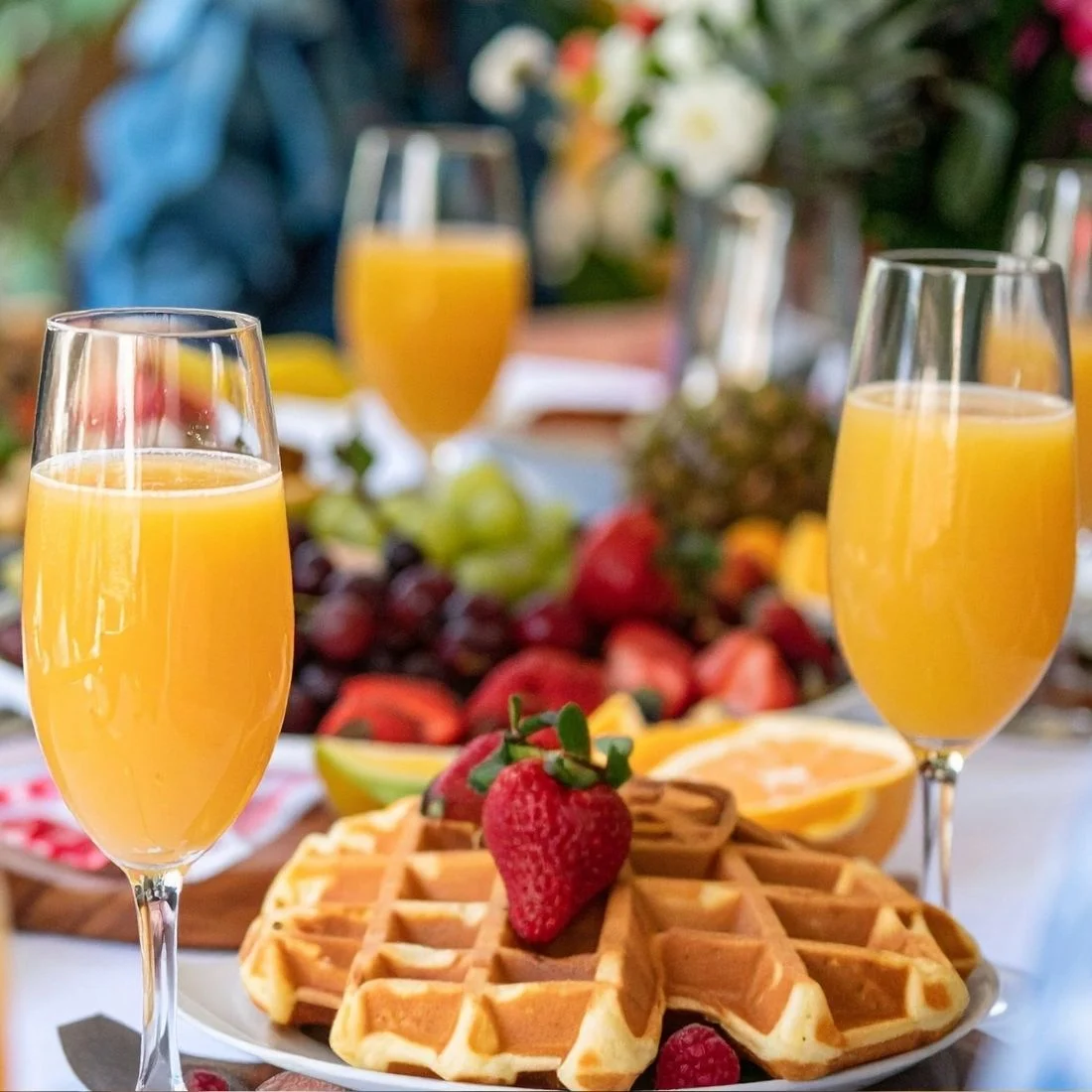 Glasses of orange juice sit on a table with fresh fruit, including strawberries, grapes, and a pineapple, alongside golden waffles topped with strawberries. The setting appears bright and inviting.