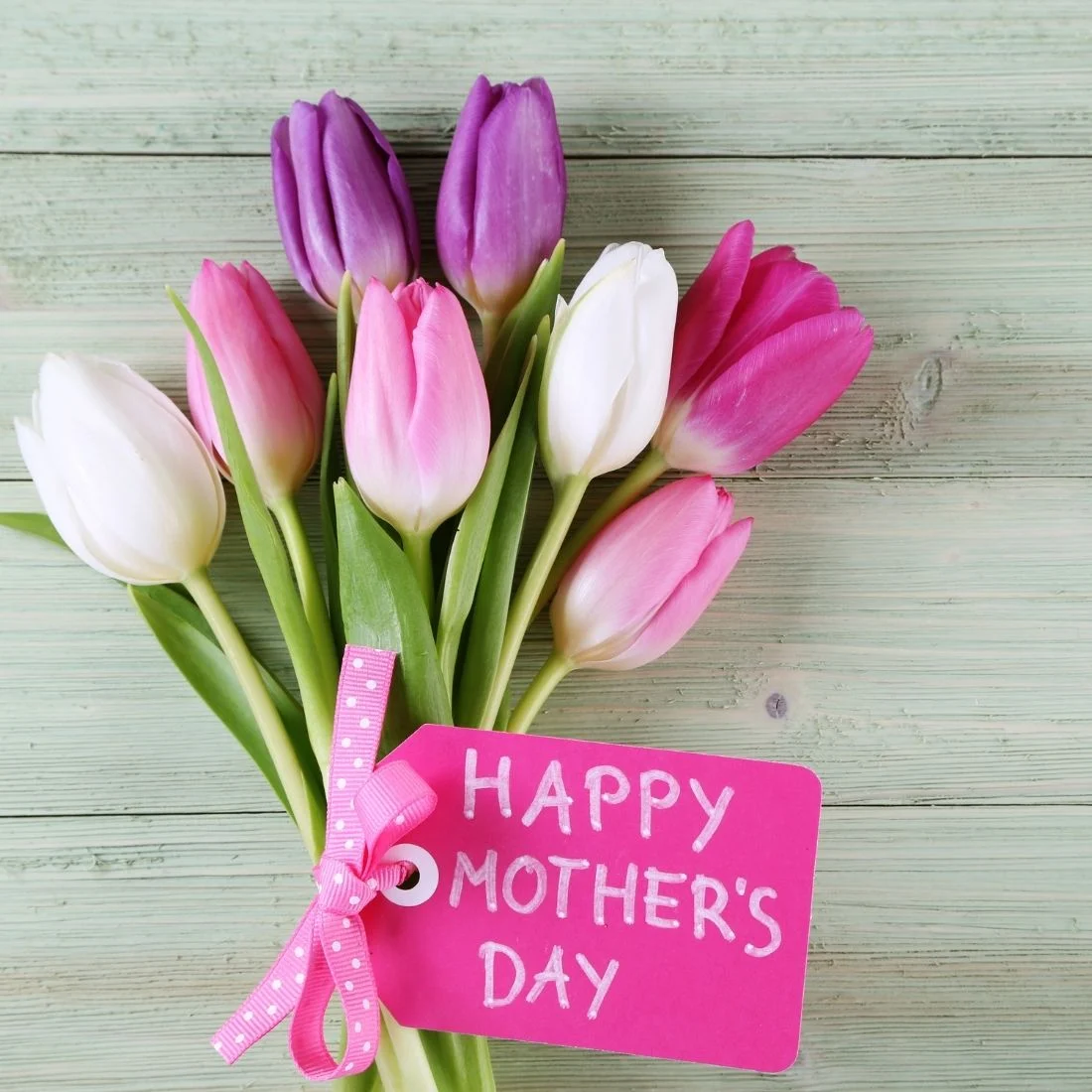 A bouquet of pink, purple, and white tulips with a pink tag that reads Happy Mothers Day, set on a light wooden surface.