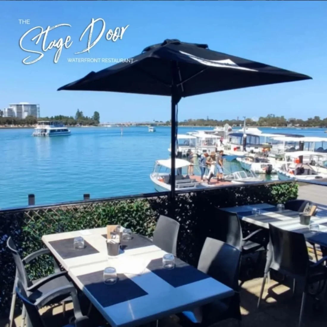 Outdoor dining area at The Stage Door Waterfront Restaurant overlooking a blue river with boats docked nearby, shaded tables and chairs, and a large black umbrella under a sunny sky.
