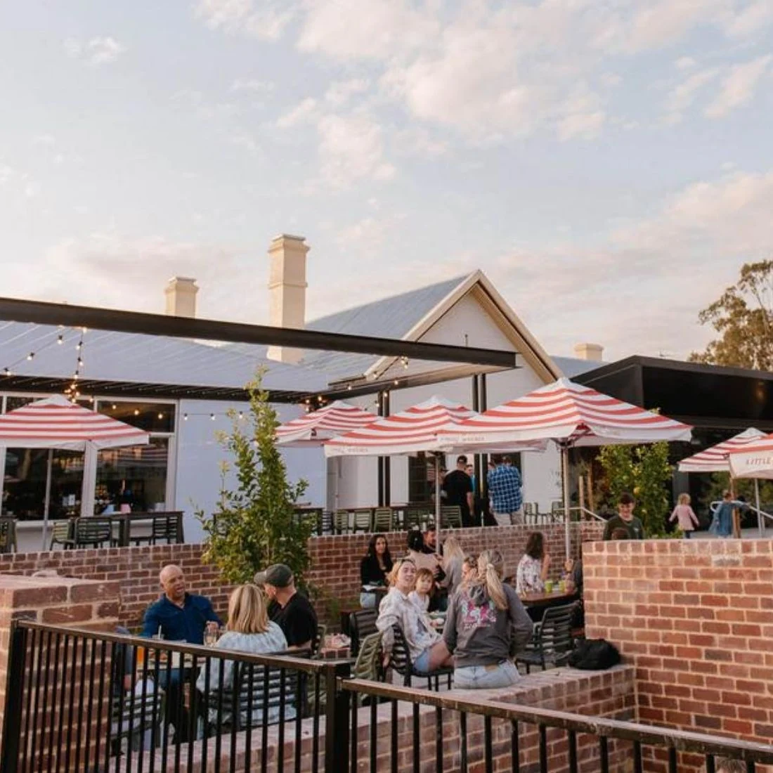 People are dining and socializing outdoors at a restaurant patio with red and white striped umbrellas, brick walls, and string lights under a partly cloudy sky.