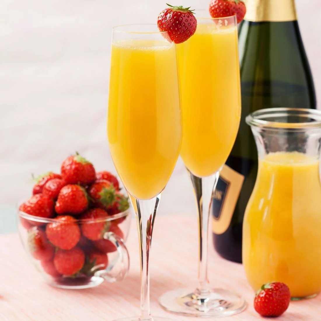 Two champagne flutes filled with orange-colored mimosa drinks, each garnished with a strawberry. A bowl of fresh strawberries, a carafe of orange juice, and a champagne bottle are in the background on a pink surface.