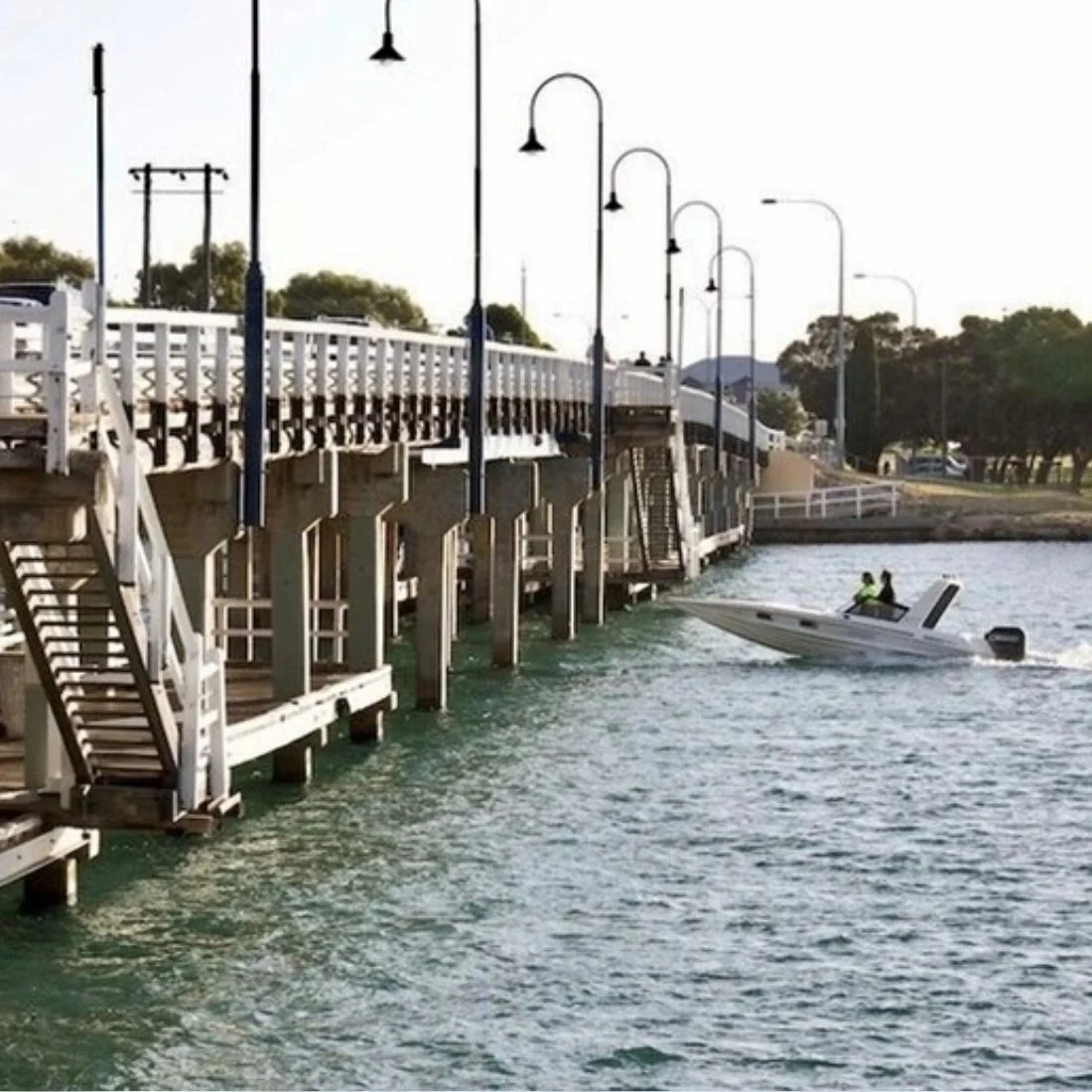 A speedboat passes under a raised section of a wooden bridge over greenish water, with two people aboard and several street lamps lining the bridge. Trees and a clear sky are in the background.