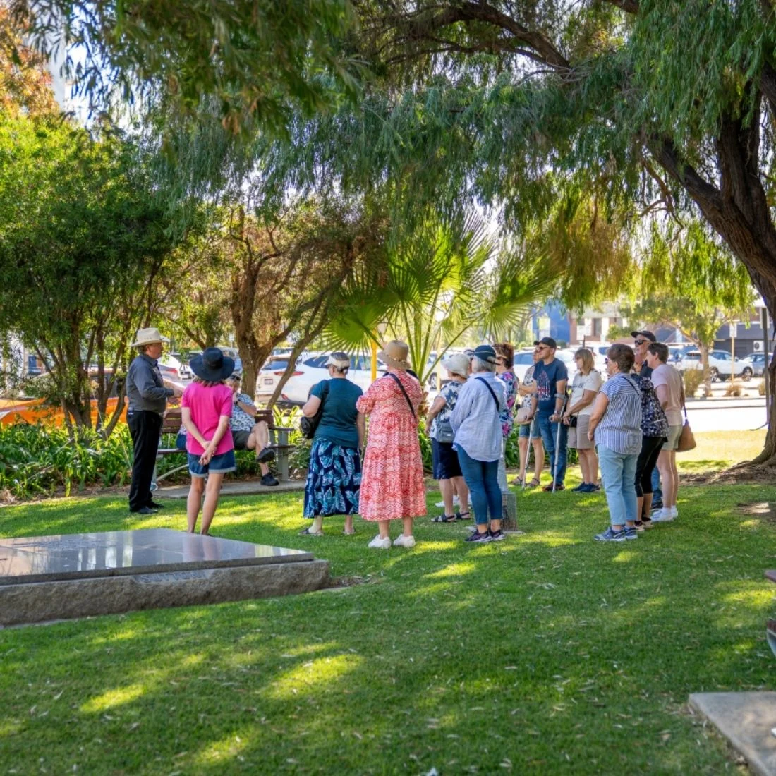 A group of people stands on grass under trees, listening to a person speaking near a gravestone in a cemetery on a sunny day. Some wear hats, and a few are looking at the speaker, while others face away.
