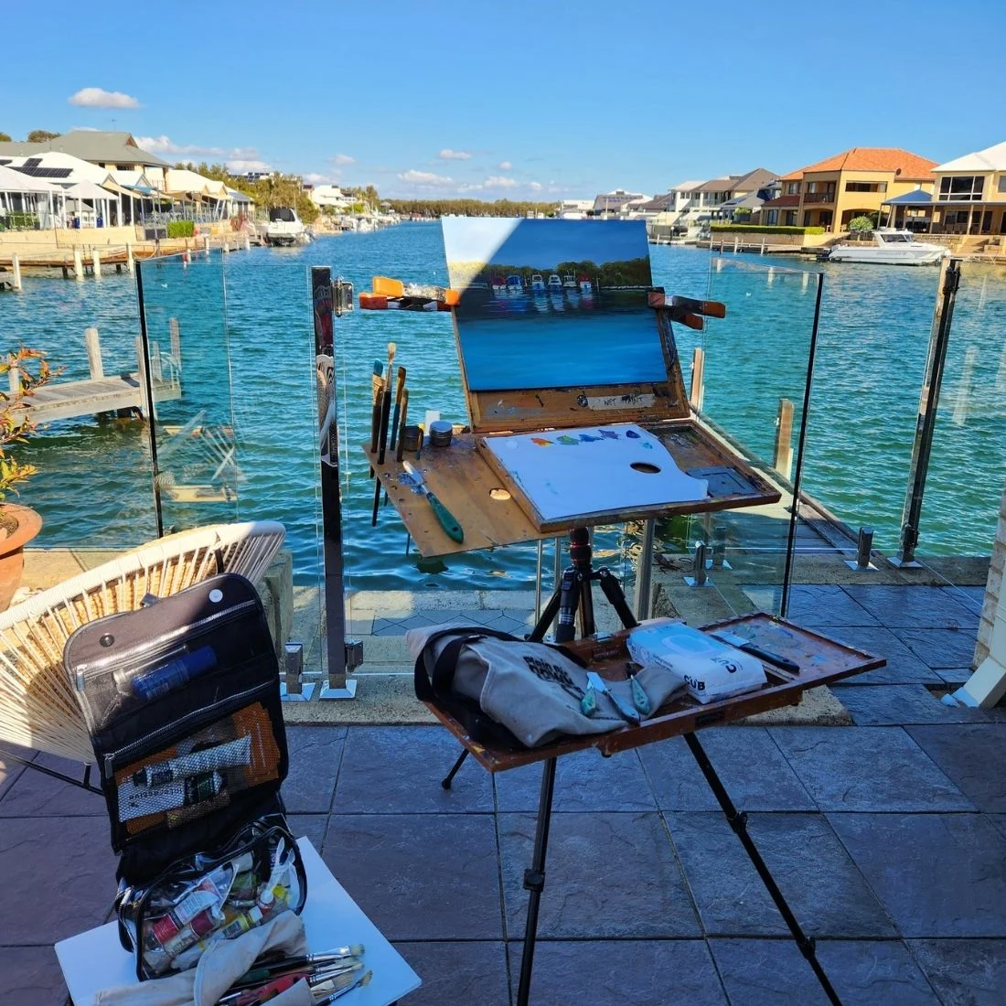 An artist’s easel and painting supplies are set up on a patio overlooking a canal with houses and boats. The canvas shows a painting in progress of the waterfront scene under a blue sky.