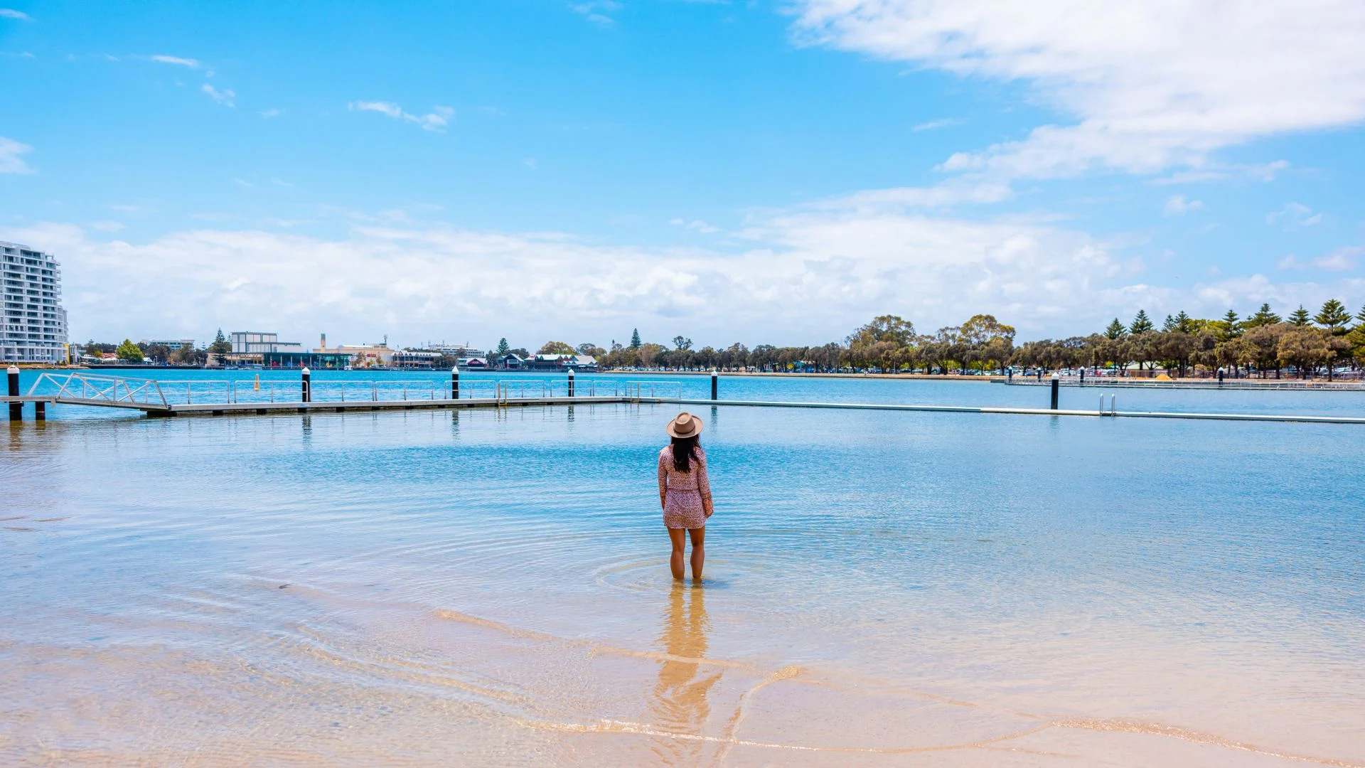 A woman stands in shallow water facing a calm blue lake with a distant shoreline of trees and buildings under a partly cloudy sky.