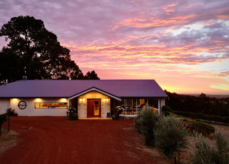 A single-story house with warm exterior lights sits on reddish soil, surrounded by greenery, under a dramatic sunset sky with pink, purple, and orange clouds. Trees frame the scene on the left.