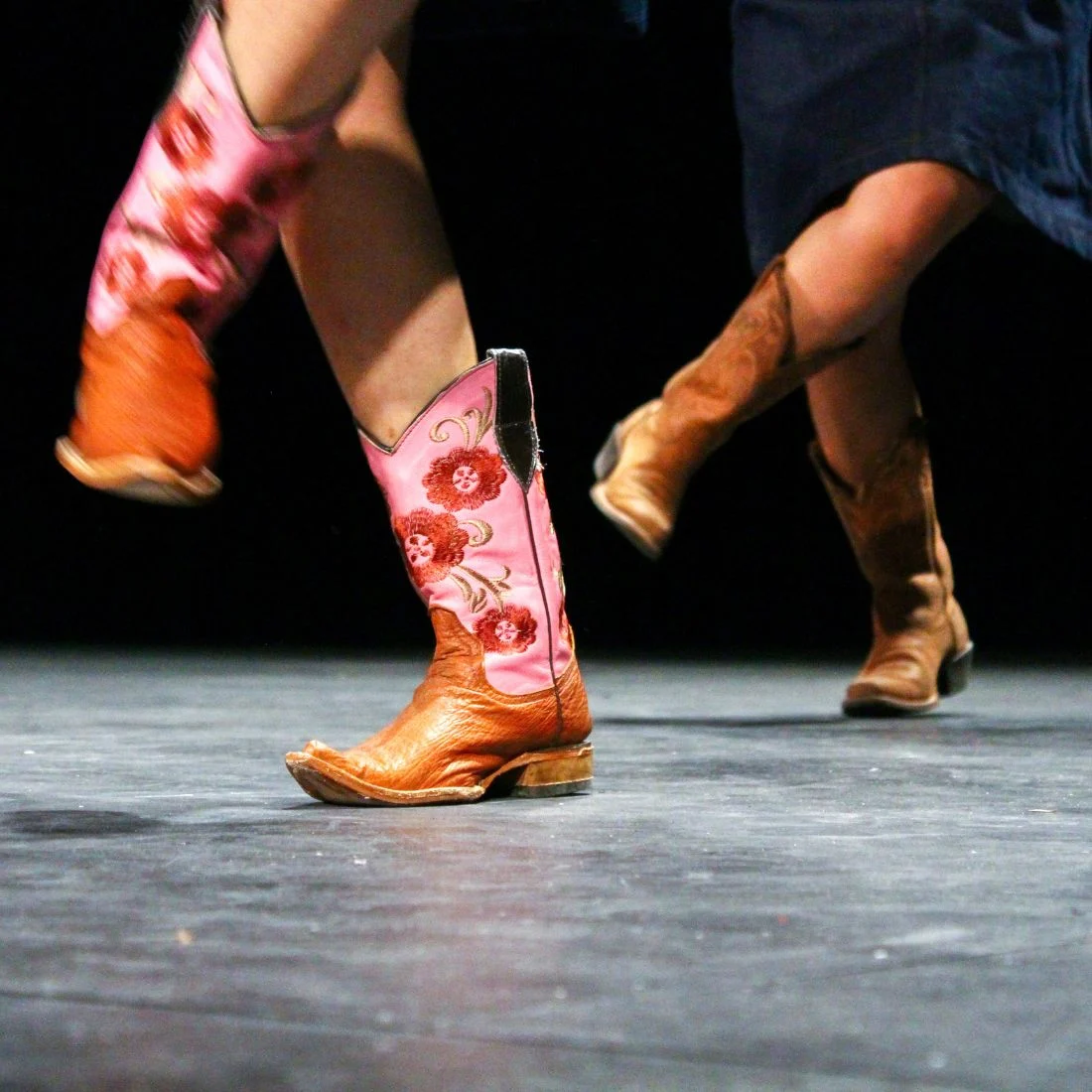 Close-up of two pairs of cowboy boots in motion, one pink with floral embroidery and one brown, on a dark stage.