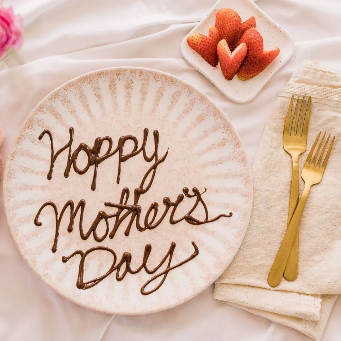 A plate with "Happy Mother's Day" written in chocolate icing. A small dish of heart-shaped strawberries and two gold forks are nearby.