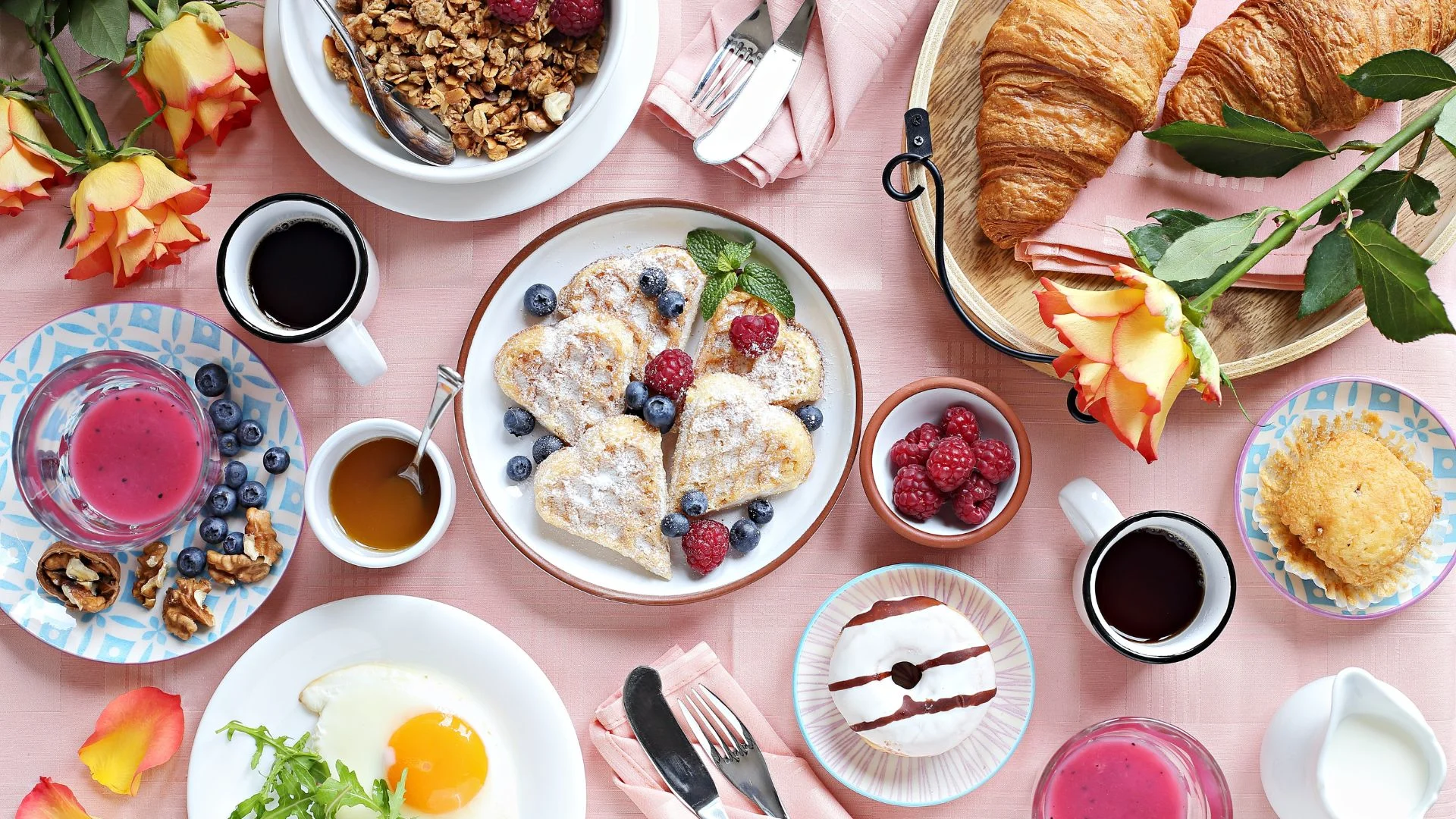 A breakfast spread with heart-shaped pancakes, croissants, coffee, juice, and roses on a pink tablecloth.