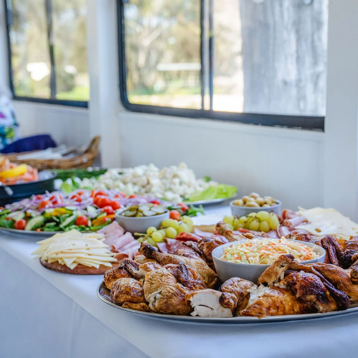 A buffet table laden with various dishes including roasted chickens, salads, cheese, ham, grapes, and coleslaw, set against a backdrop of windows with outdoor scenery.
