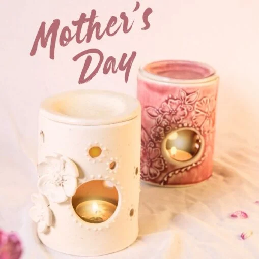 Two decorative ceramic candle holders, one white with floral designs and one pink with embossed flowers, each holding a lit tealight. Mothers Day is written above in large script.