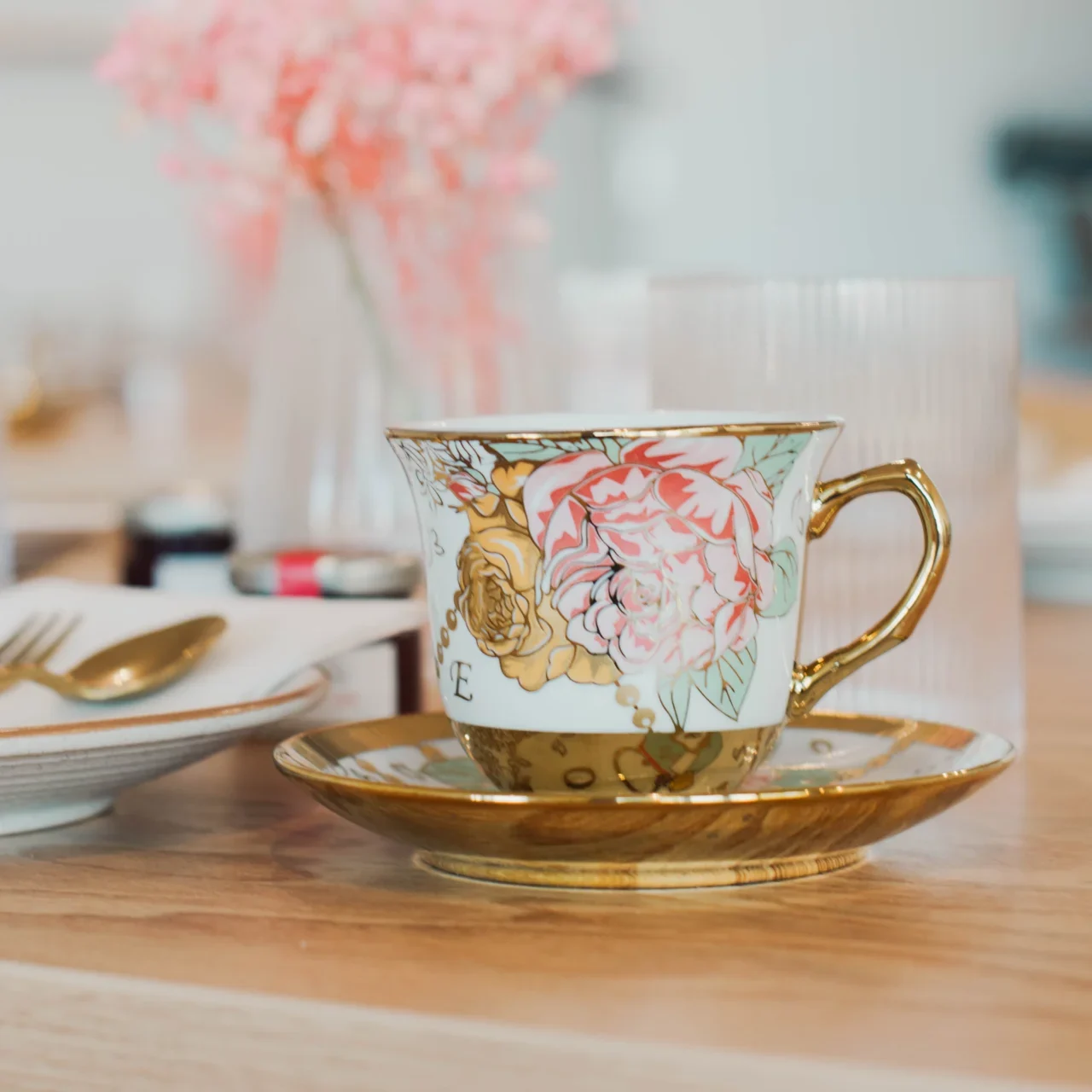 A decorative teacup with gold accents and floral patterns sits on a matching saucer on a wooden table, with a plate, utensils, and a blurred pink flower arrangement in the background.