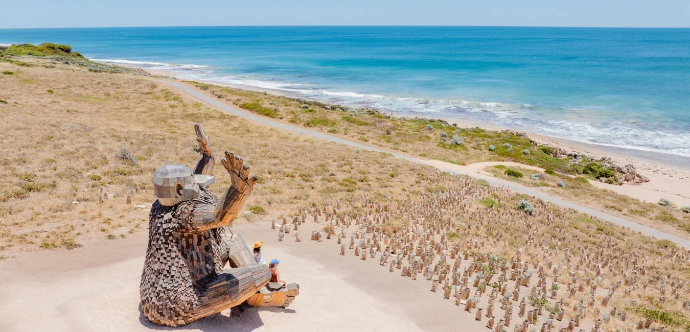 A large wooden sculpture of a seated figure overlooks a beach with the ocean in the background and a path winding through dry grass and small rock formations.