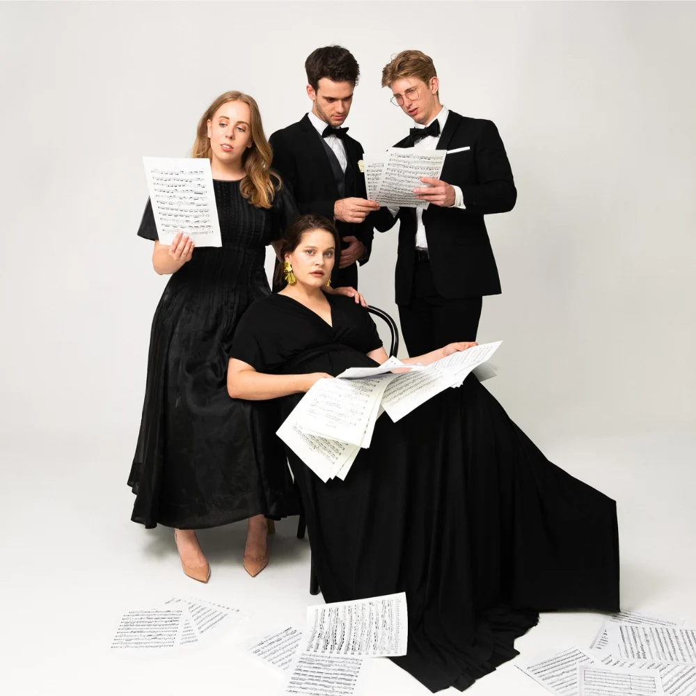 Four people in formal black attire hold and read sheet music. One woman sits with music sheets on her lap and floor, while the other three stand around her, all looking focused against a plain white background.